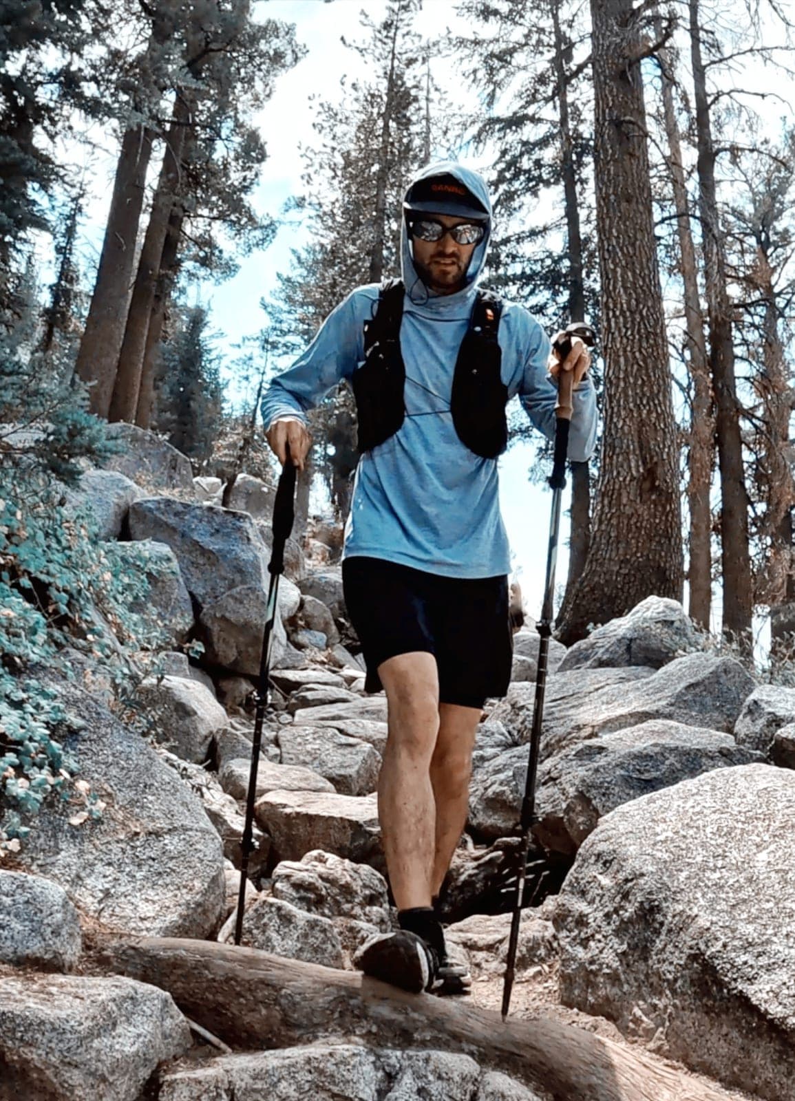 A runner in a blue sun hoody climbs a granite staircase through pines, a pole in each hand