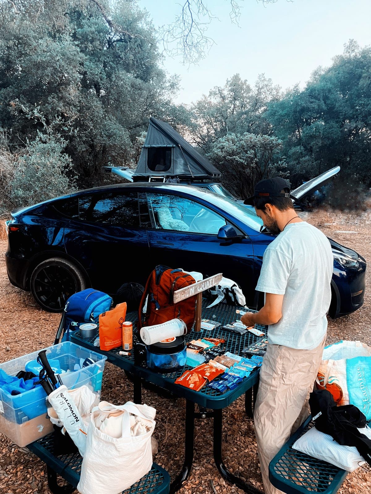A crew table at a trailhead laid out with gels, bars and drink mix beside drop bags and a car with a rooftop tent